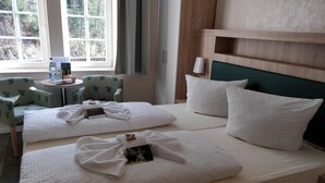 A hotel room with two beds, a wooden headboard, a window with a view of trees, a chair, and a small table with a water bottle and a menu. - Gaststätte & Pension Lichtenhainer Wasserfall (Sebnitz)