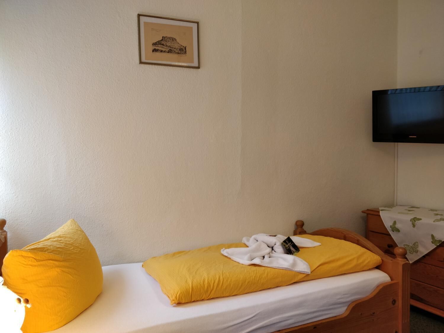 A single bed with a yellow pillow and a white blanket, a wooden nightstand with a television above it, and a framed picture on the wall.