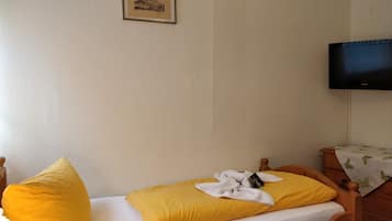 A single bed with a yellow pillow and a white blanket, a wooden nightstand with a television above it, and a framed picture on the wall.