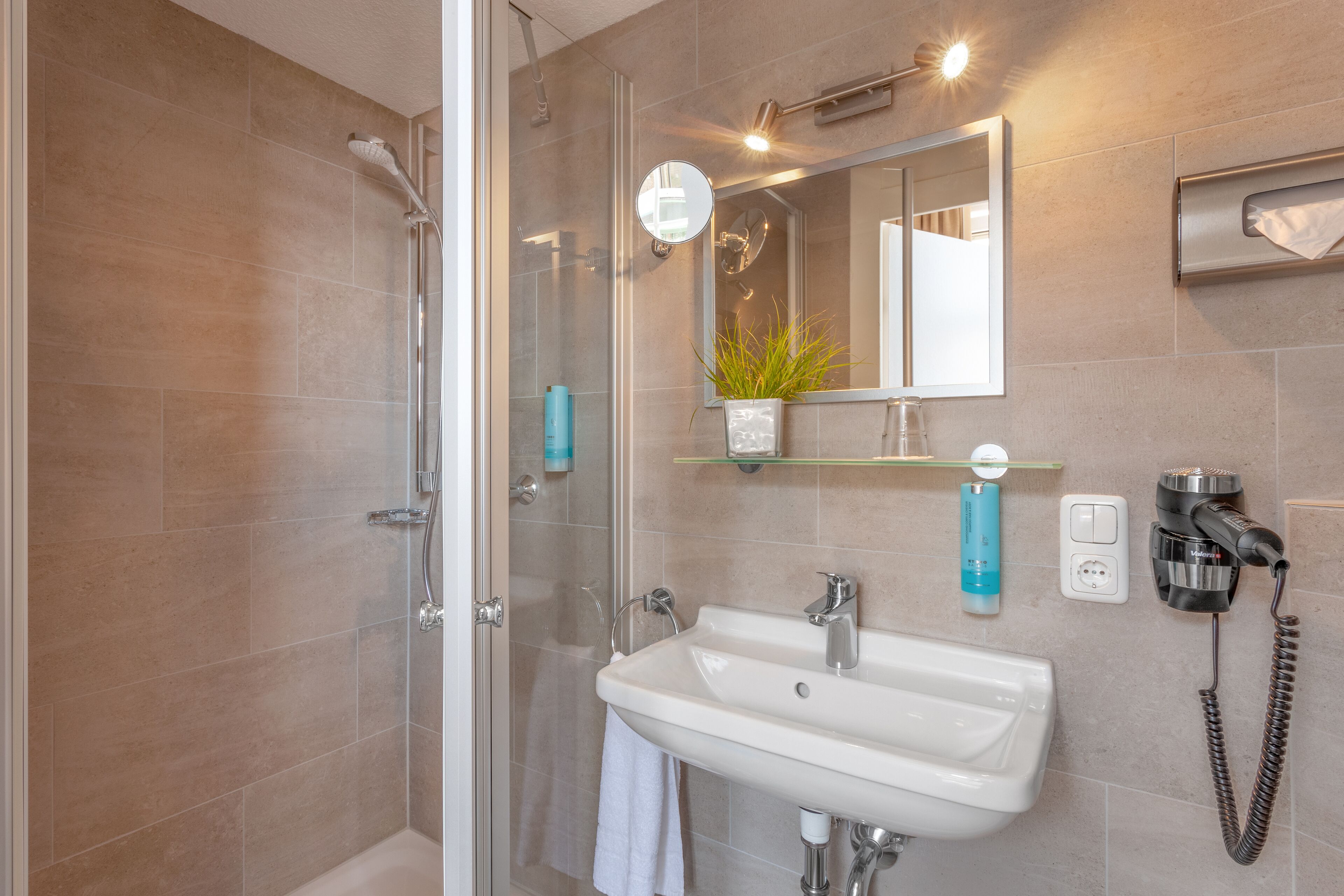 A bathroom with a glass shower enclosure, a white sink, a mirror, and a hairdryer on a wall-mounted charging station.