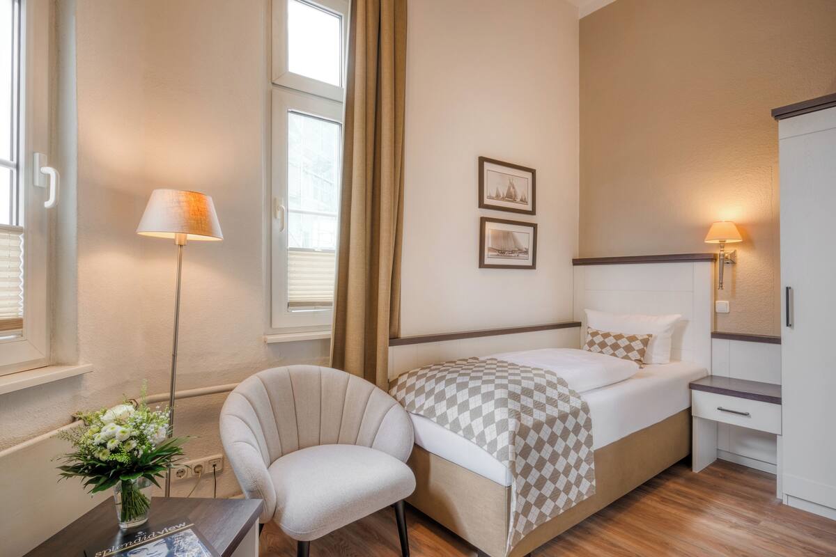 Una habitación de hotel con cama, una silla, un escritorio con lámpara y un jarrón con flores.