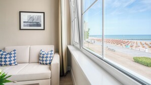 Une pièce dotée d’une grande fenêtre donnant sur une plage, d’un canapé blanc agrémenté de coussins à motifs et d’un tableau encadré accroché au mur.