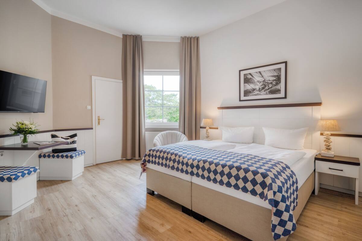 Habitación de hotel con cama, mesitas de noche, televisor, ventana con cortinas y un cuadro en la pared.