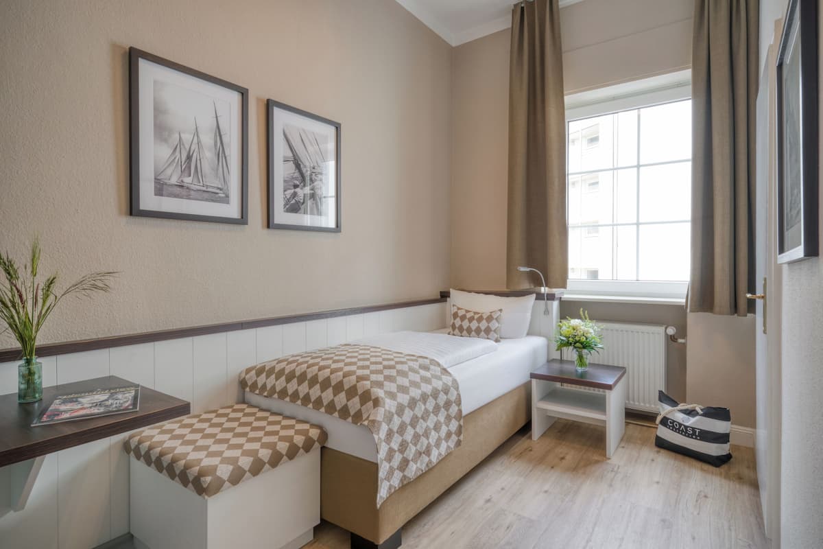 Habitación de hotel con cama, mesita de noche, ventana con cortinas y dos cuadros en la pared.