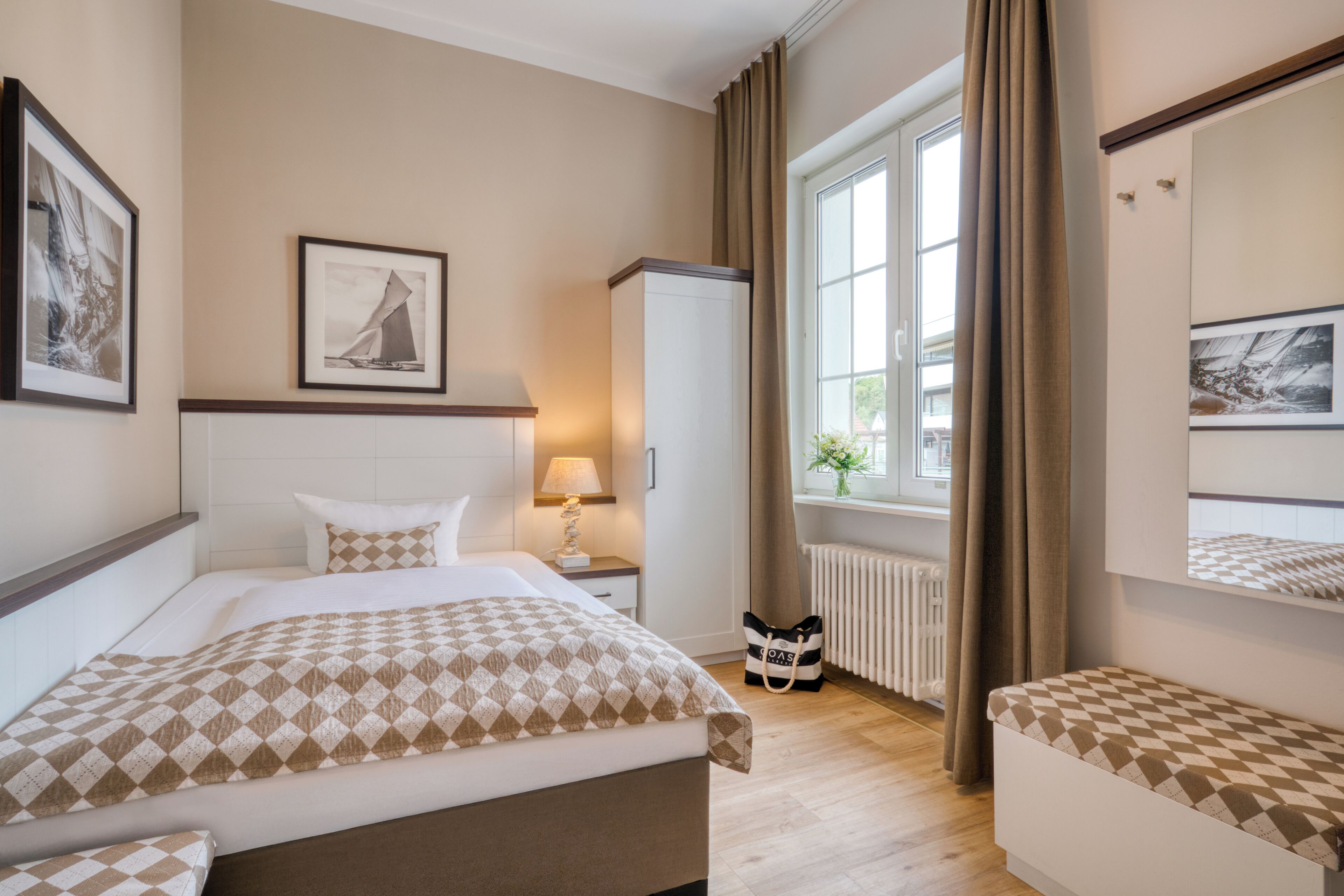 Ein Hotelzimmer mit einem Bett, einem Nachttisch, einer Lampe, einem Schrank, einem Fenster mit Vorhängen und einer Wand mit gerahmten Bildern.