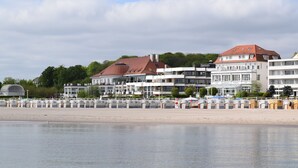 Un hôtel situé au bord de l’eau, composé de plusieurs bâtiments, avec une plage en premier plan et une rangée de cabanes de plage numérotées.