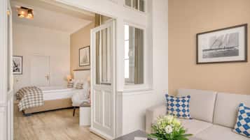 Quarto bem iluminado com uma cama, um sofá e uma mesa de centro com flores. Há uma porta que dá para outro cômodo, e um quadro na parede.