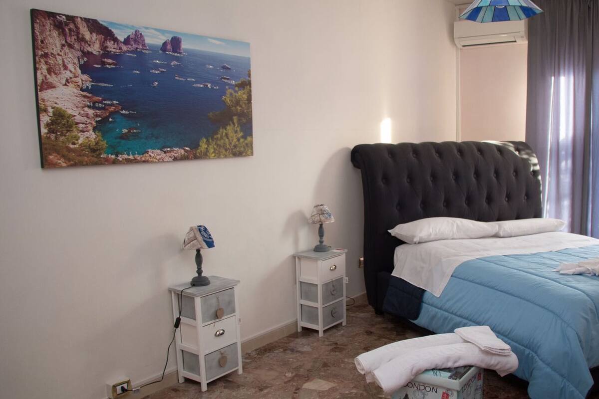 A bedroom with a large painting of a coastal landscape, a black upholstered headboard, a blue bedspread, and a white bedside table with a lamp.