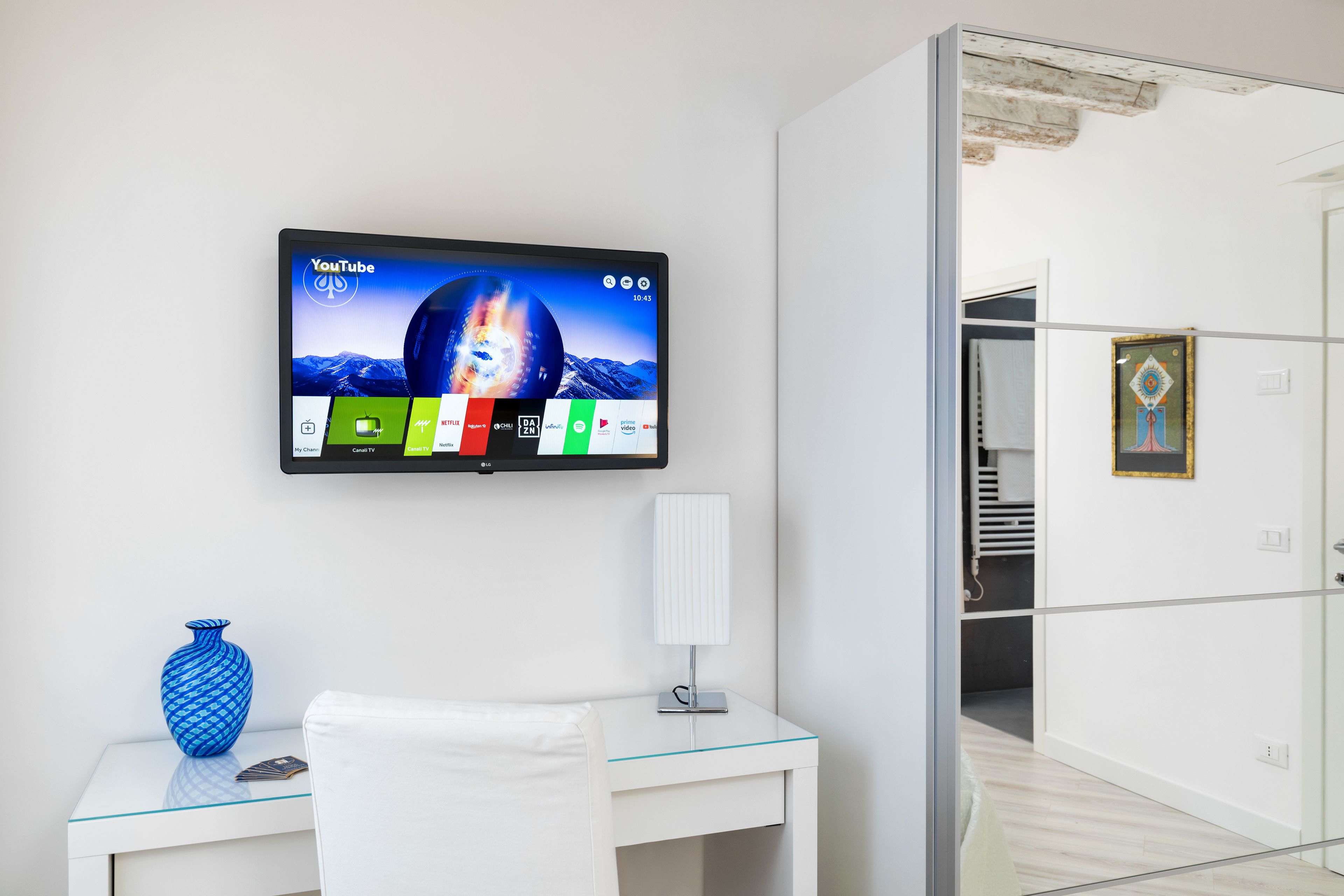 Ein modernes Hotelzimmer mit einem an der Wand befestigten Fernseher, der YouTube anzeigt, einem Glastisch mit weißem Stuhl, einer blauen Vase und einer weißen Lampe.