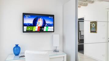 Ein modernes Hotelzimmer mit einem an der Wand befestigten Fernseher, der YouTube anzeigt, einem Glastisch mit weißem Stuhl, einer blauen Vase und einer weißen Lampe.