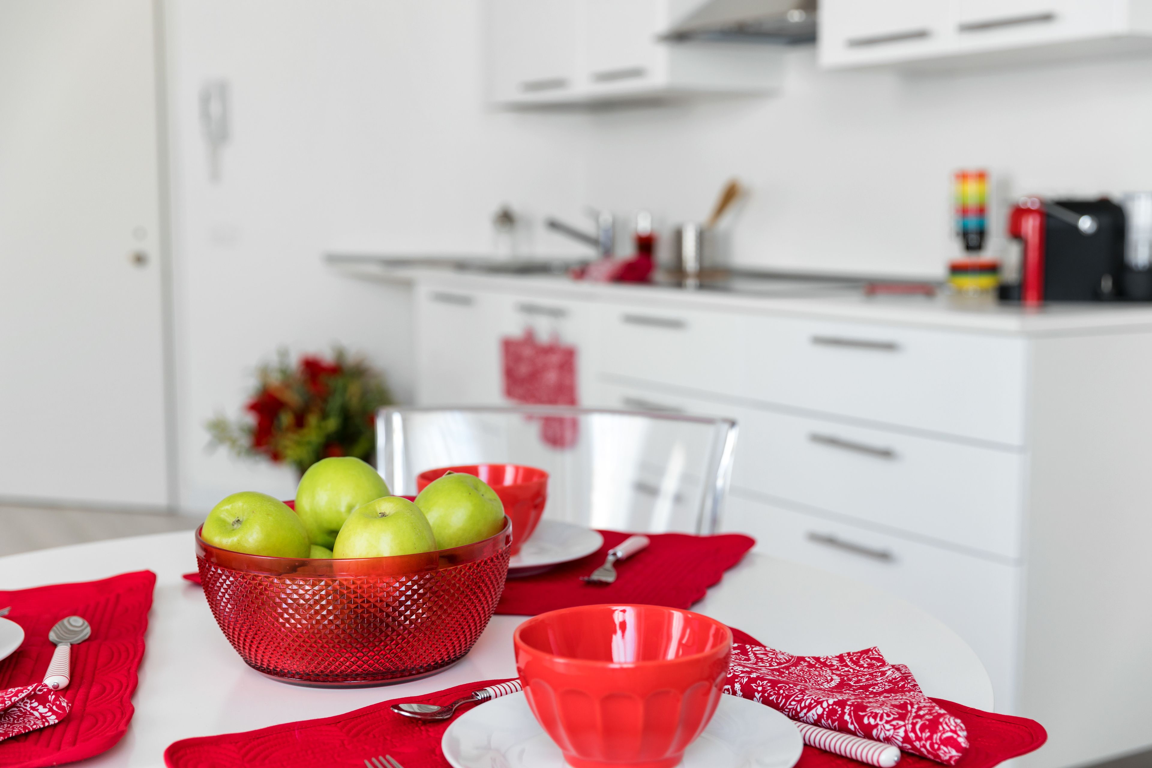 A kitchen with white cabinets, a red bowl with apples, and a red cup on the table.