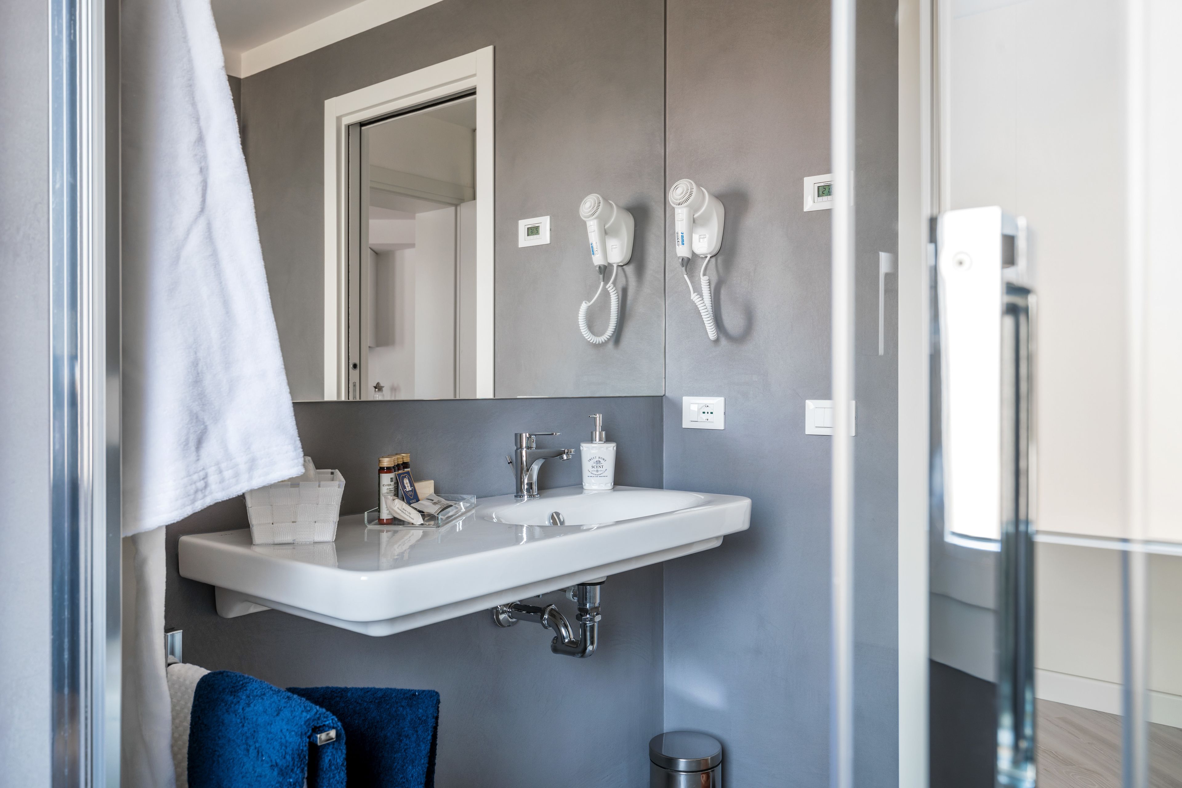 A modern bathroom with a white sink, two hair dryers mounted on the wall, a mirror, and a blue towel.