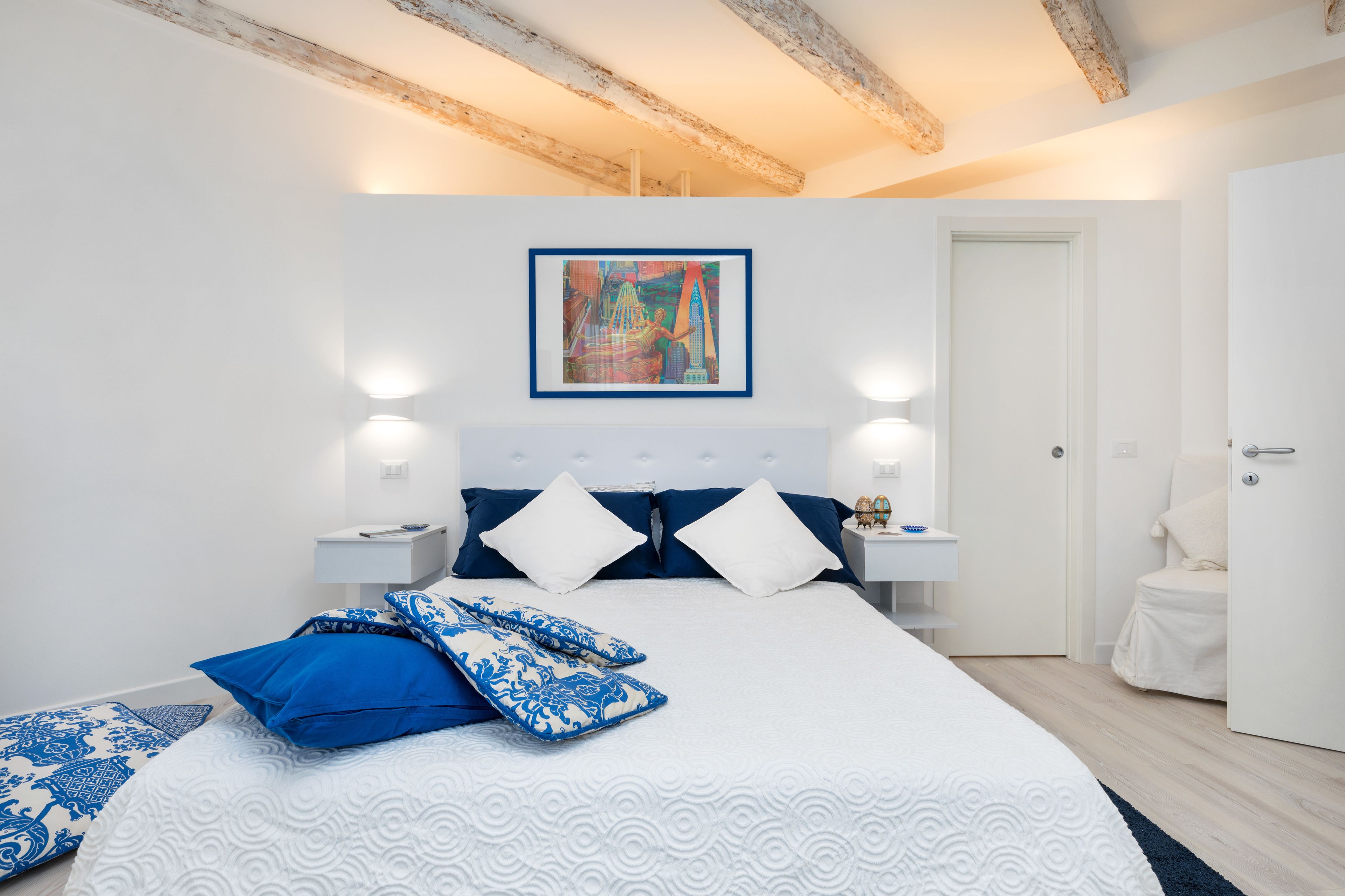 Ein Schlafzimmer mit einem großen Bett, weißen Wänden, einer Holzbalkendecke, einem gerahmten Gemälde, Nachttischlampen und einer weißen Tür.