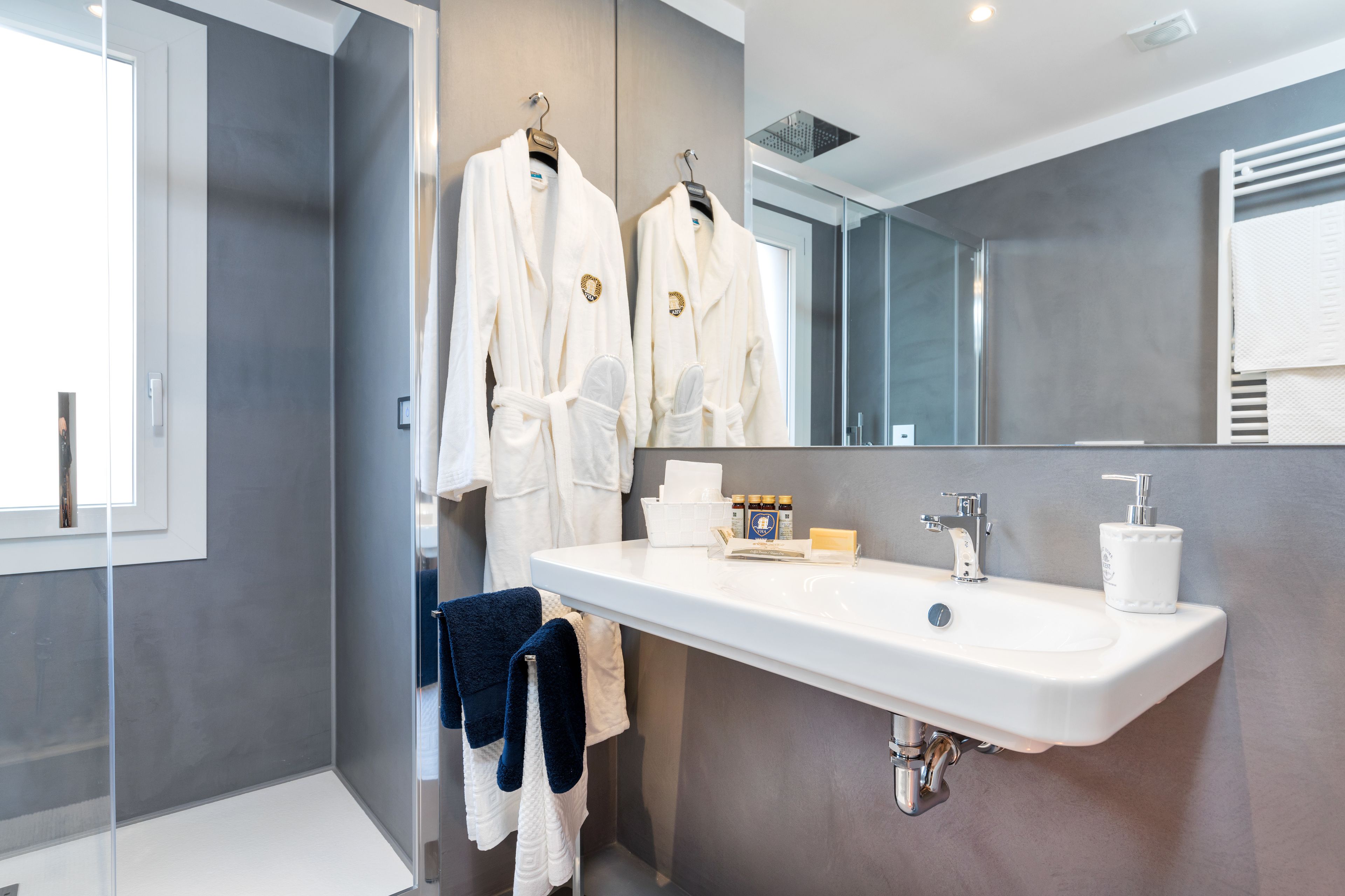 Un baño moderno con un espejo grande, un lavabo blanco y dos albornoces blancos colgados en un perchero.
