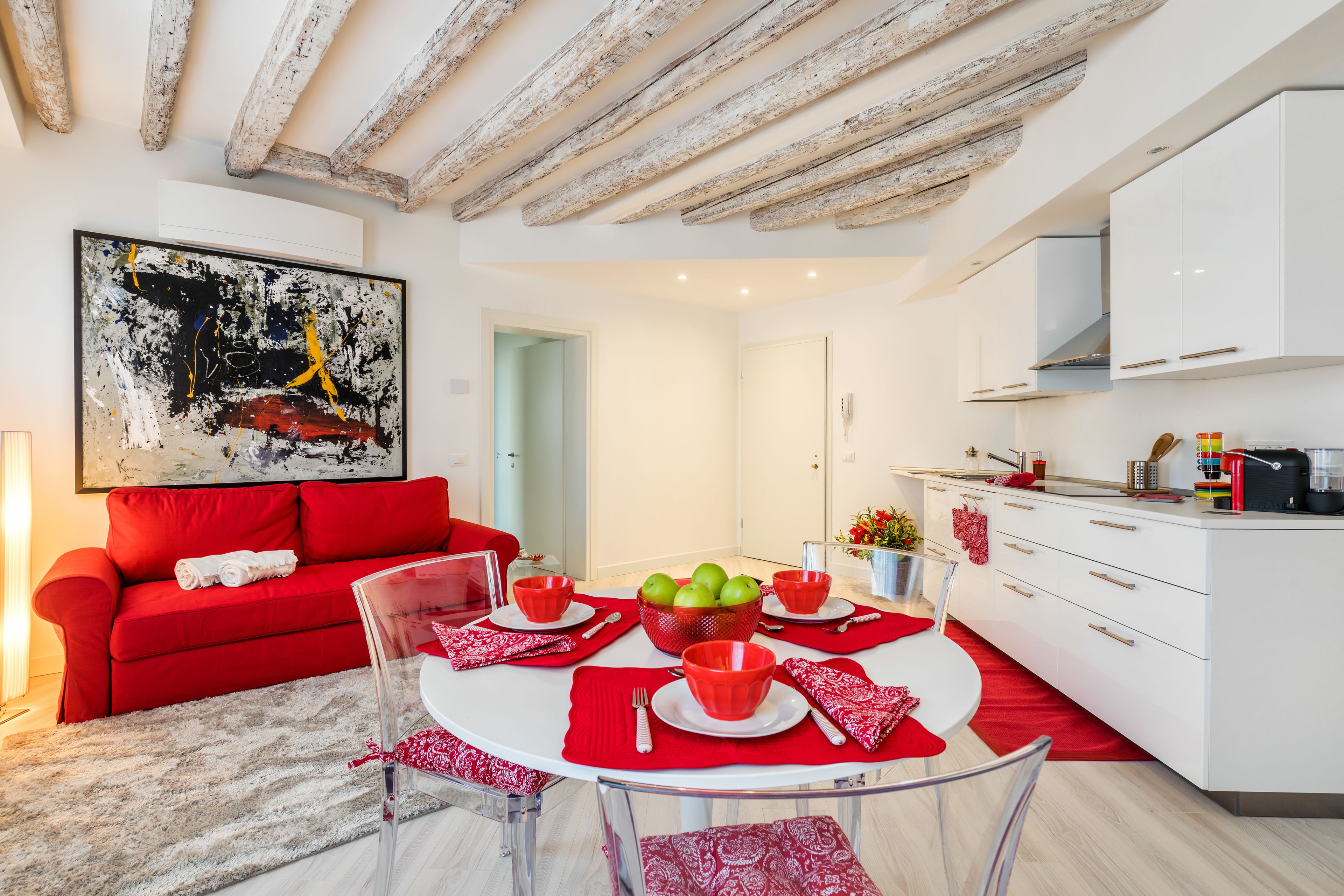 A modern living space with a red sofa, a glass dining table with red placemats, and a kitchen area with white cabinets.