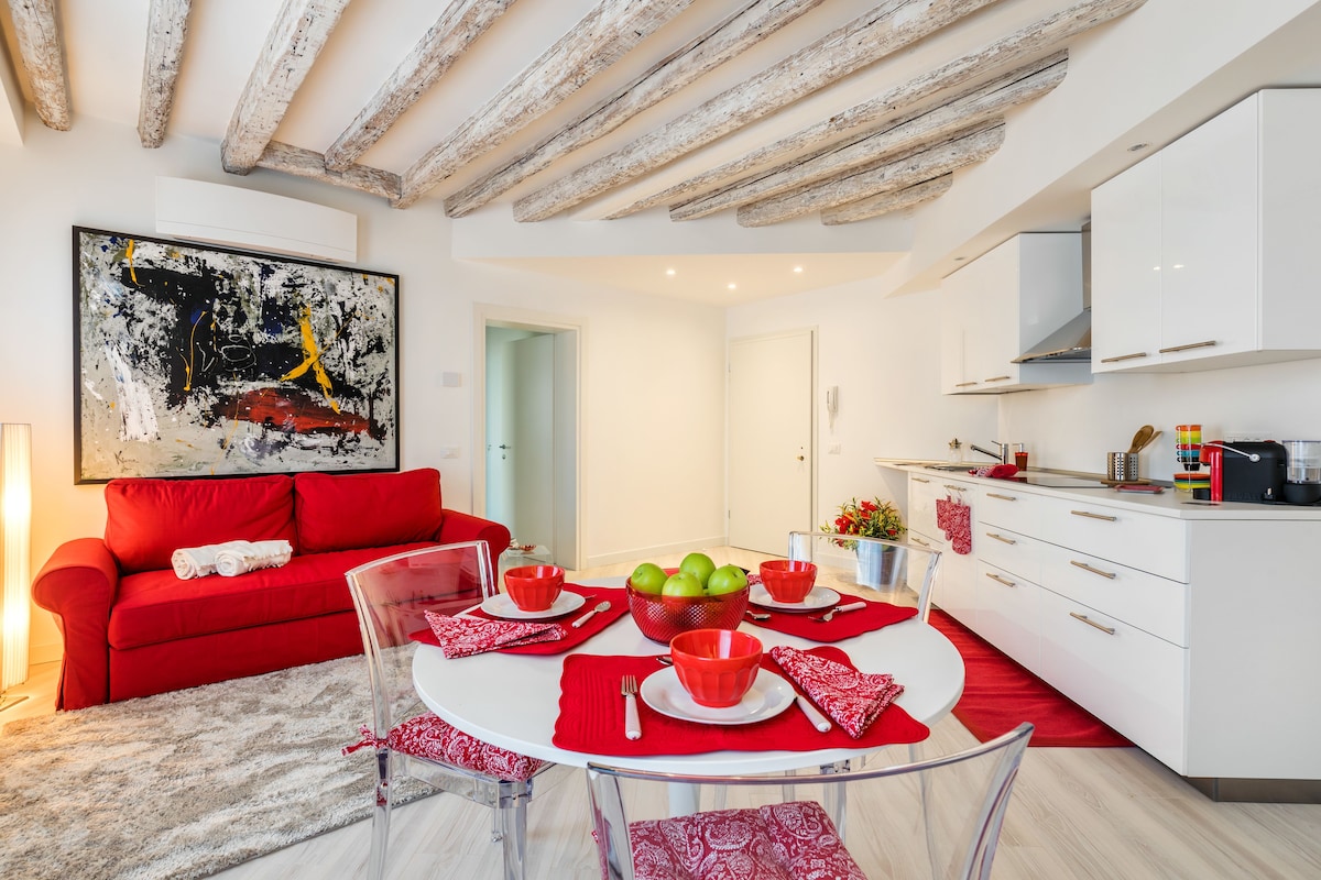A modern living space with a red sofa, a glass dining table with red placemats, and a kitchen area with white cabinets.