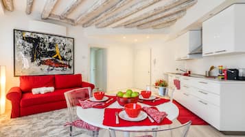 A modern living space with a red sofa, a glass dining table with red placemats, and a kitchen area with white cabinets.