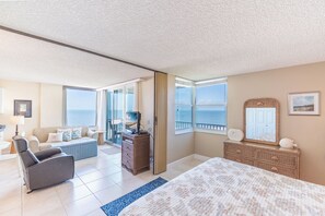 A spacious bedroom with a large bed, a television on a stand, a dresser, and a view of the sea through the windows.