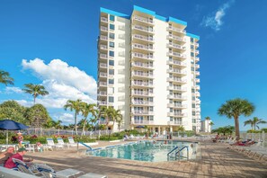 A high-rise hotel with a swimming pool, palm trees, and a clear blue sky.