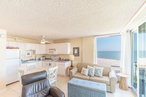 Une cuisine moderne avec des meubles blancs, un coin repas avec une table et des chaises, un canapé avec des coussins et une grande fenêtre donnant sur la plage.