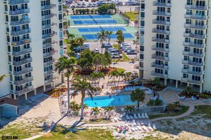 A high-rise hotel with multiple balconies, a tennis court, a swimming pool, and a parking area.