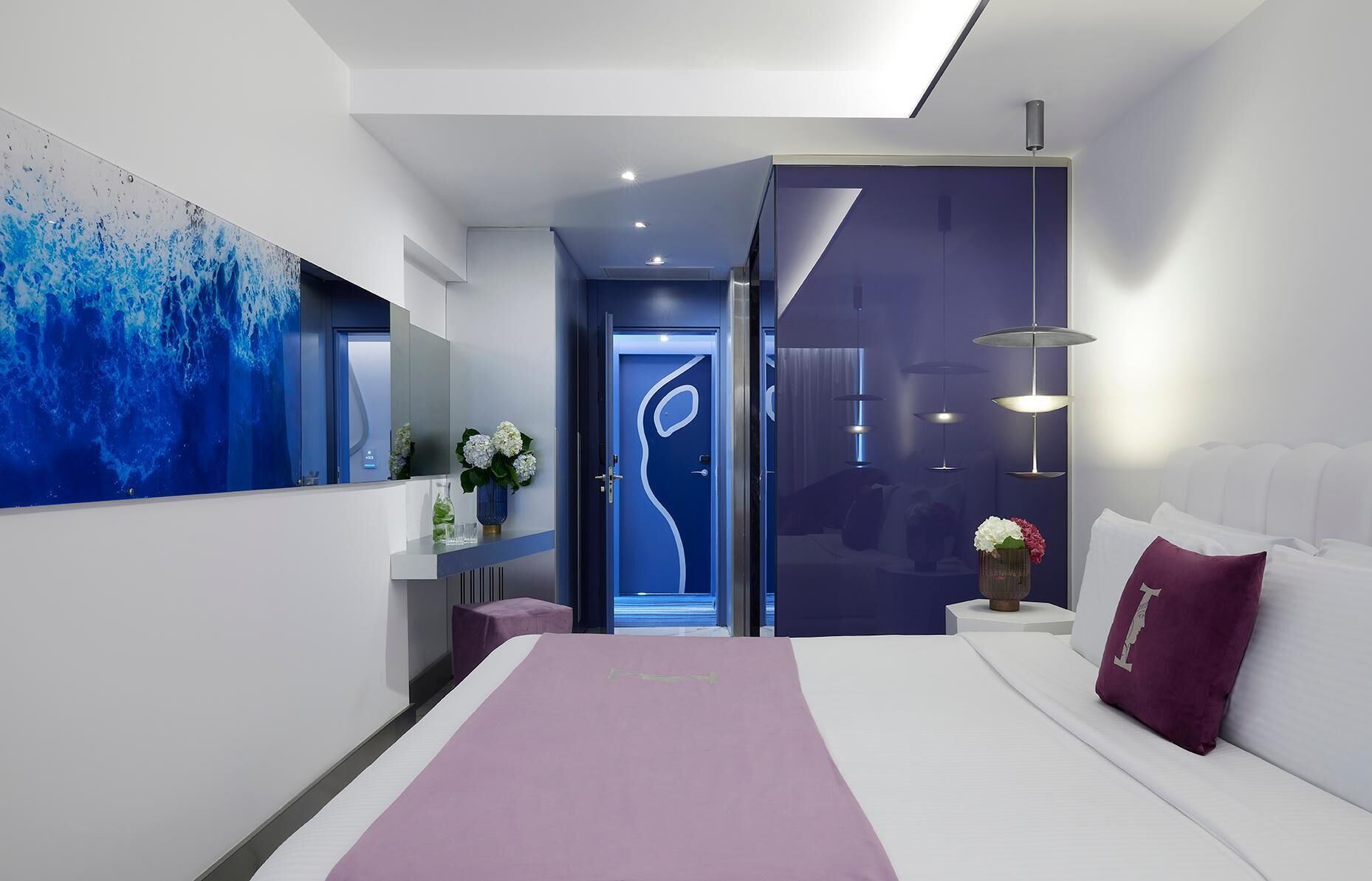 Un dormitorio moderno con una cama grande, una mesita de noche, un florero con flores y un cuadro abstracto azul en la pared.
