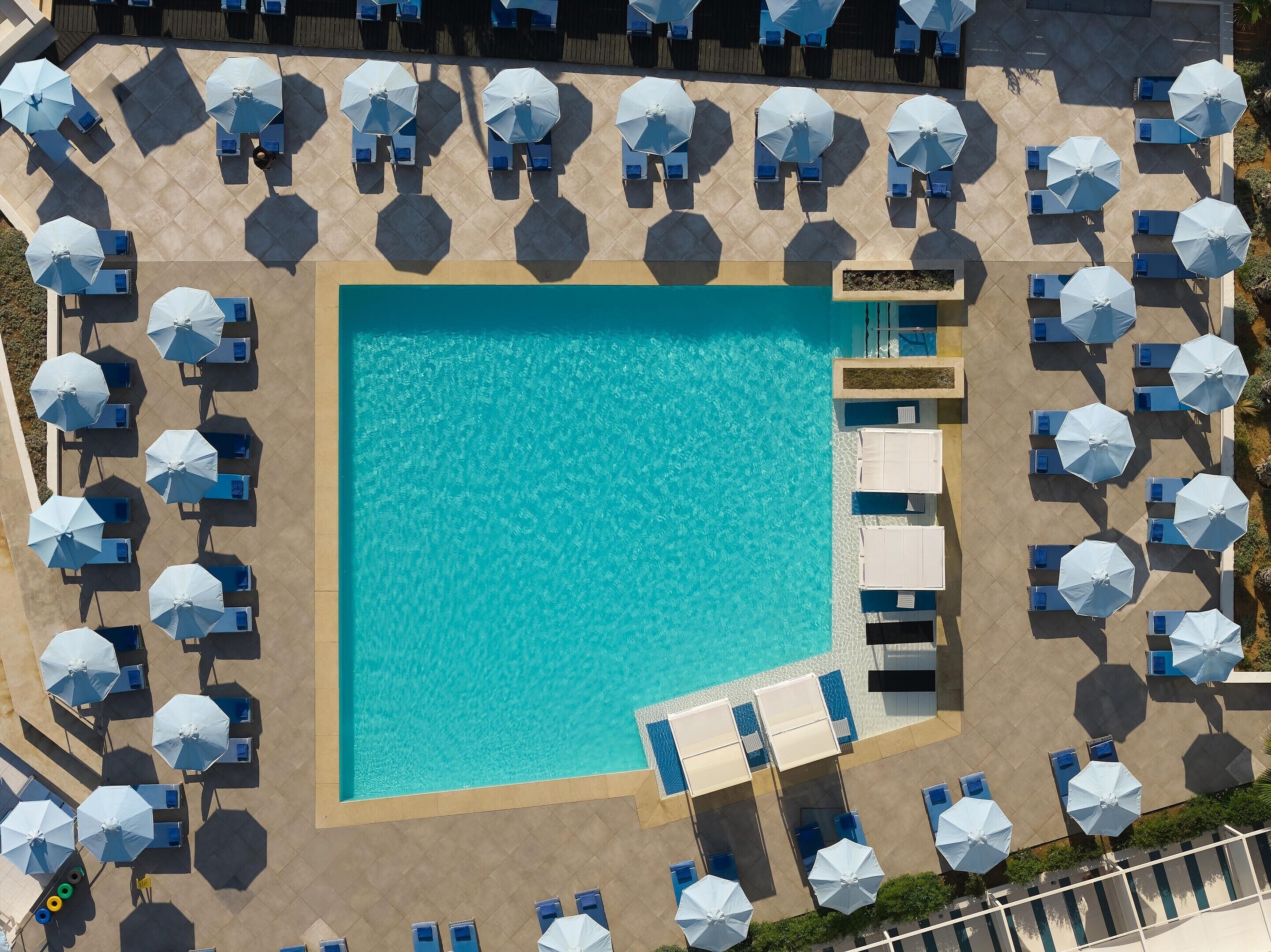 An aerial view of a hotel with a central swimming pool surrounded by multiple rows of sunbeds and umbrellas.