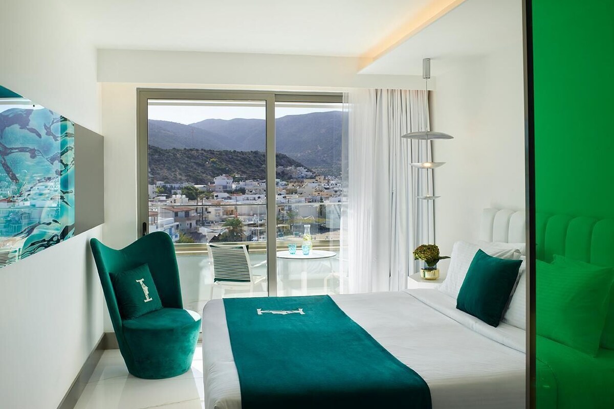 Een moderne hotelkamer met een groot bed, een groene fauteuil, een balkon met uitzicht op de stad, en een groot raam.