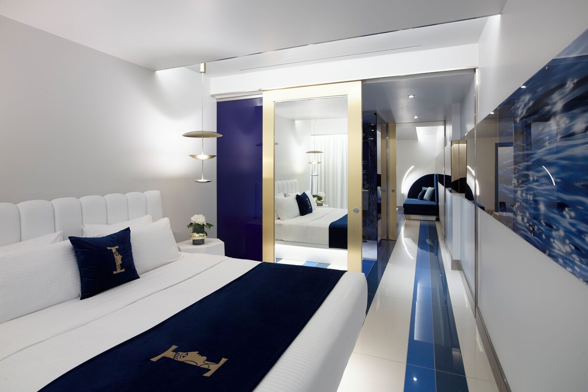 Een moderne hotelkamer met een groot bed, een nachtkastje met een vaas en een spiegel met een blauwe marmeren achterwand.