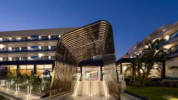 Una entrada de hotel moderna con un arco metálico distintivo, un edificio de varios pisos con balcones y un jardín bien cuidado.