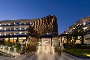 L’entrée d’un hôtel moderne se distingue par une arche métallique originale, un immeuble à plusieurs étages doté de balcons et un jardin bien entretenu.