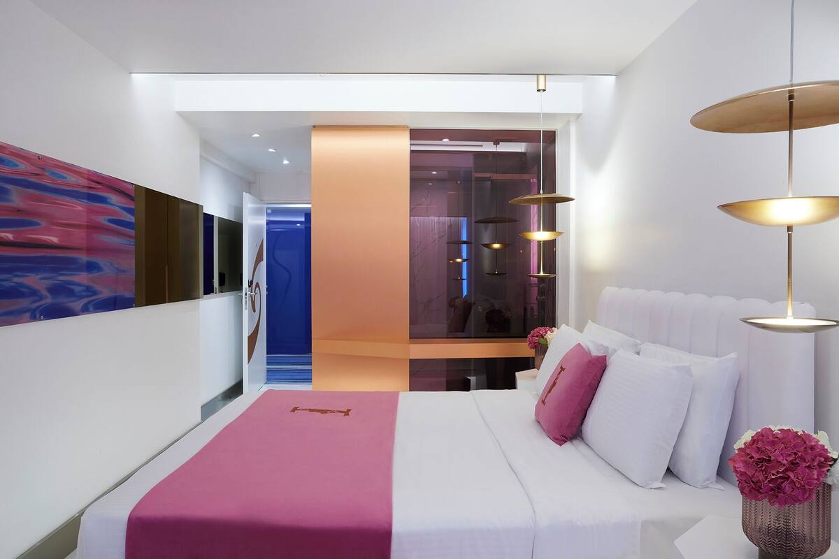 Une chambre d’hôtel moderne avec un grand lit, un mur d’accent rose et une vue sur le couloir.