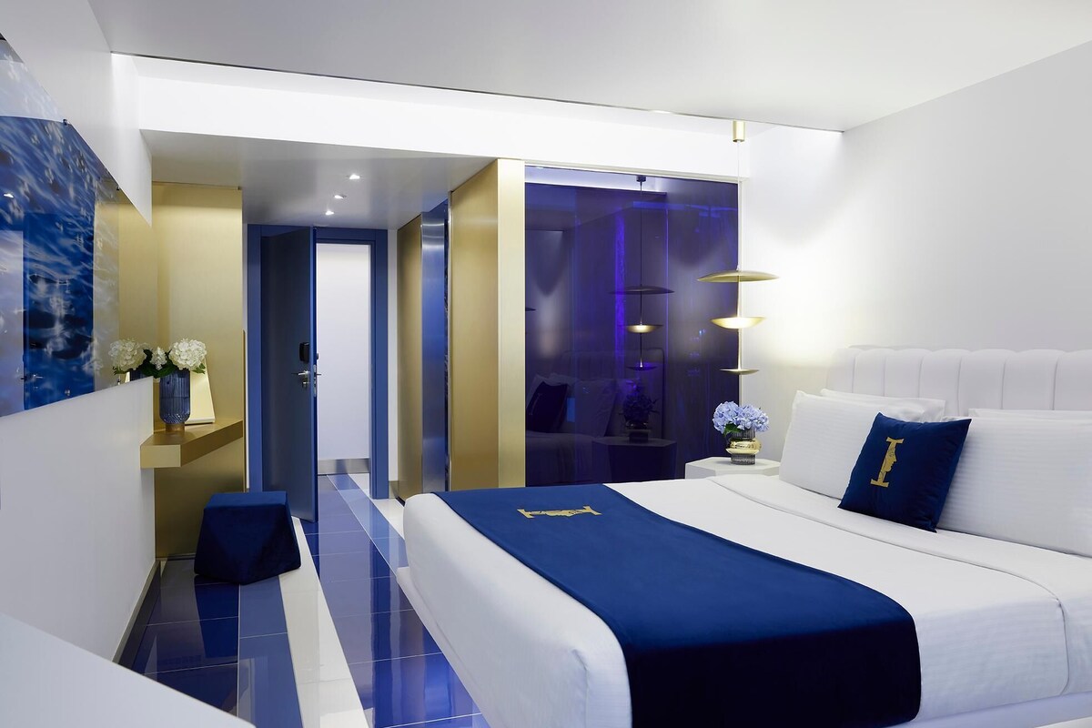 Een moderne hotelkamer met een groot bed, een nachtkastje en een bijbehorend tafeltje. Er is een deur die naar een andere kamer leidt.