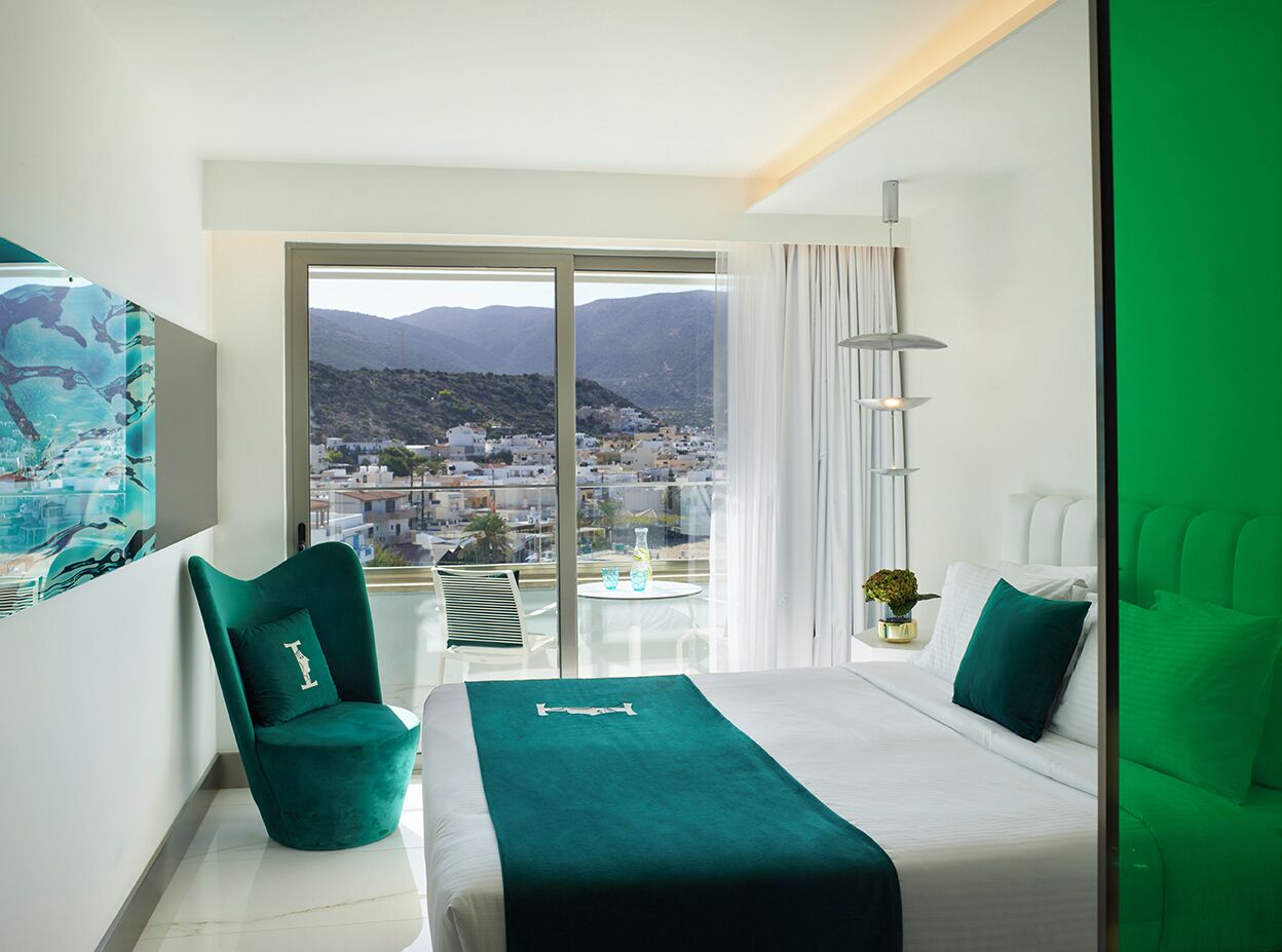 A modern hotel room with a large balcony, a bed with a teal bedspread, a green armchair, and a view of a cityscape.