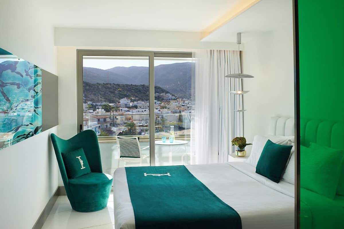 A modern hotel room with a large balcony, a bed with a teal bedspread, a green armchair, and a view of a cityscape.
