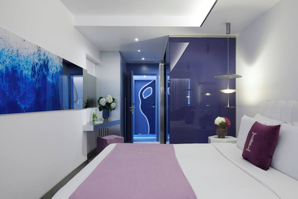 Une chambre d’hôtel moderne avec un grand lit, une table de chevet avec une lampe, un téléviseur fixé au mur et une salle de bain visible à travers une porte ouverte.
