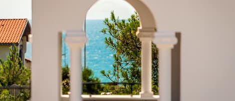 A view through an arched window with a coastal landscape, including trees and a building with a red-tiled roof.