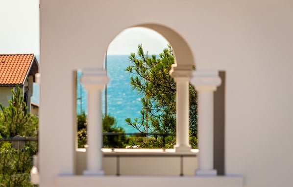 A view through an arched window with a coastal landscape, including trees and a building with a red-tiled roof.