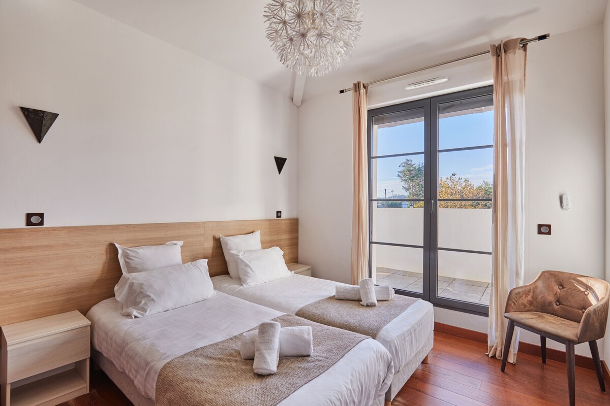 A hotel room with two beds, a wooden headboard, a chandelier, a balcony with curtains, and a chair.