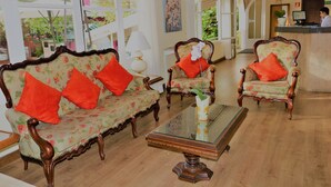 Un espace d’accueil comprenant un comptoir de réception en bois, deux fauteuils à motifs floraux avec des coussins rouges, une table basse en verre et une grande fenêtre donnant sur l’extérieur.