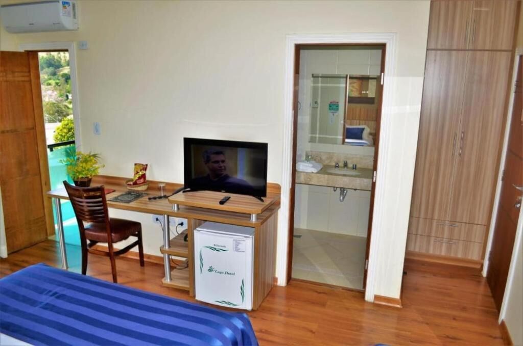 A compact room with a wooden desk, a television, a chair, a small refrigerator, and a bed with a blue bedspread.