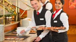 A hotel staff member serving food at a buffet counter with a glass display case and stacked plates in the background.