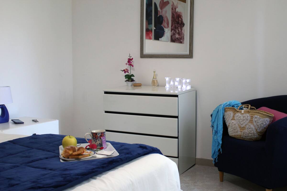 A bedroom with a bed, a dresser, a chair, a framed picture on the wall, and a tray with breakfast items.