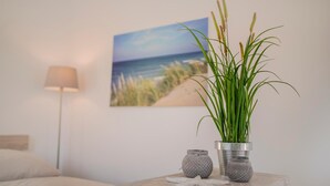 A bedroom with a bed, a nightstand with a lamp, a potted plant, and a painting of a beach scene. - BUTZ home (Scharbeutz)