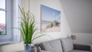 A cozy living room with a grey sofa, a small table with decorative items, a potted plant, and a beach-themed painting on the wall. - BUTZ home (Scharbeutz)