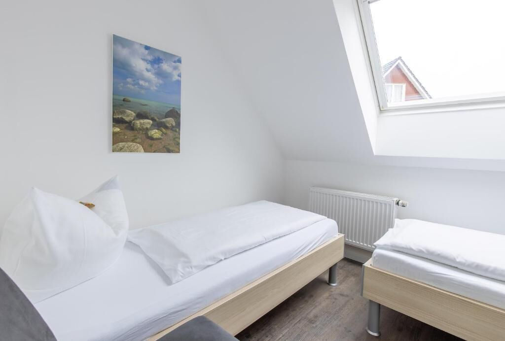 A small, single-bed room with a white bed, a white pillow, a white radiator, and a painting of a landscape on the wall.
