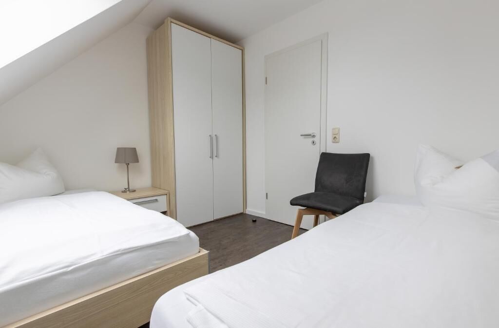 A hotel room with two beds, a wardrobe, a chair, and a bedside table with a lamp.