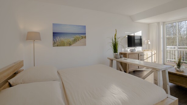 A bedroom with a large bed, a desk with a TV, a lamp, and a window with a view of the beach. - BUTZ home (Scharbeutz)
