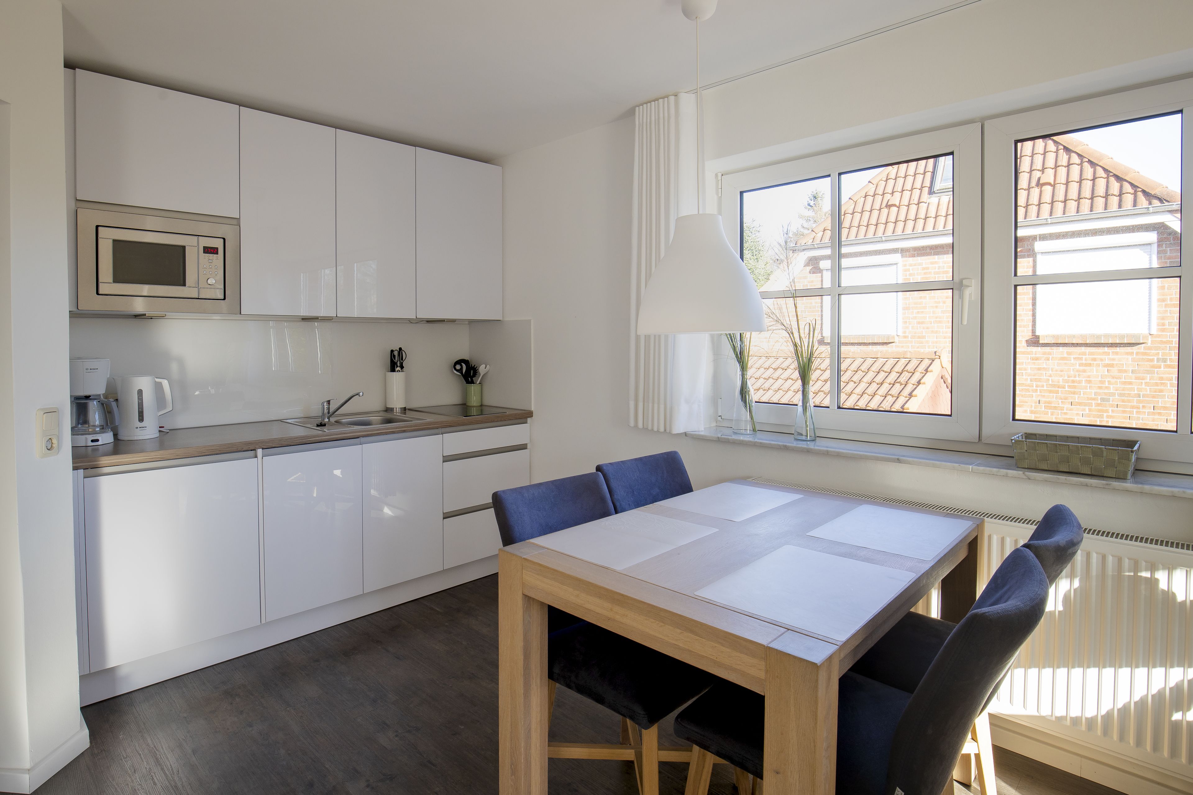 A modern kitchen with white cabinets, a microwave, and a coffee maker. There's a dining table with chairs and a window with a view of a brick building.