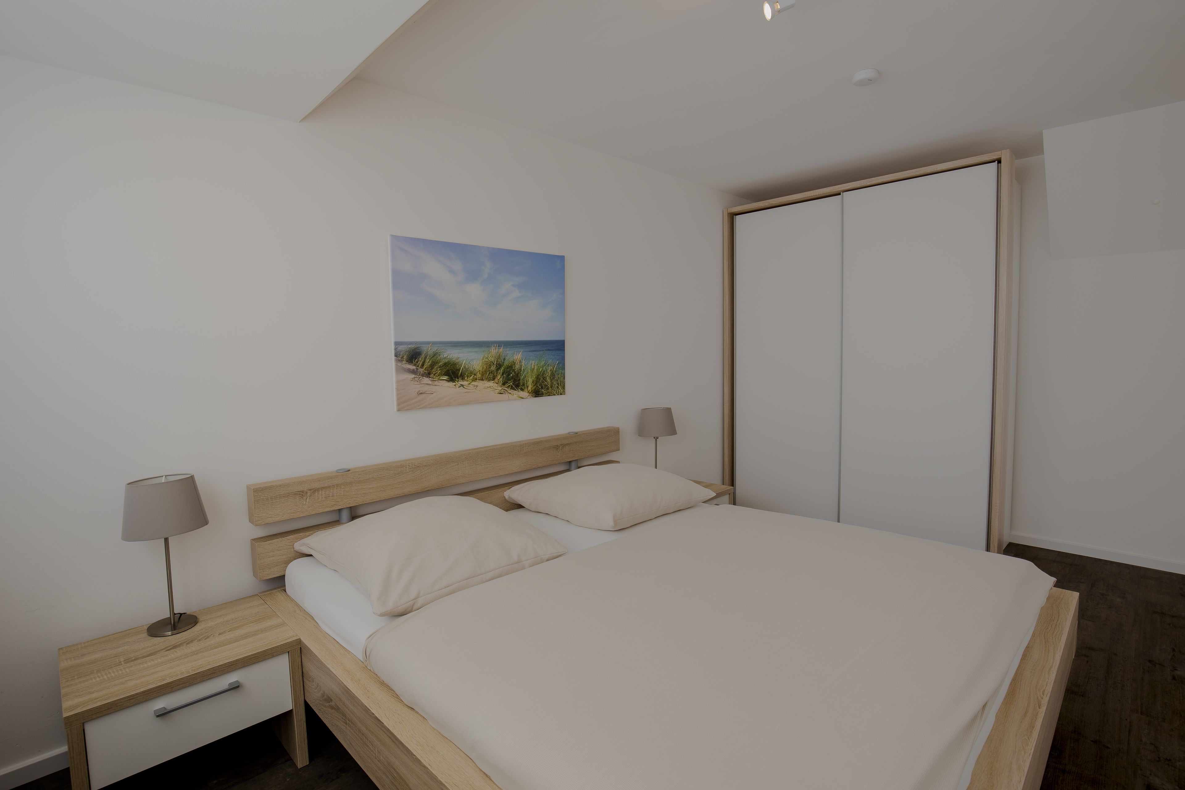 Une chambre à coucher comprenant un lit, des tables de chevet, une lampe, une armoire et un tableau sur le thème de la plage accroché au mur.